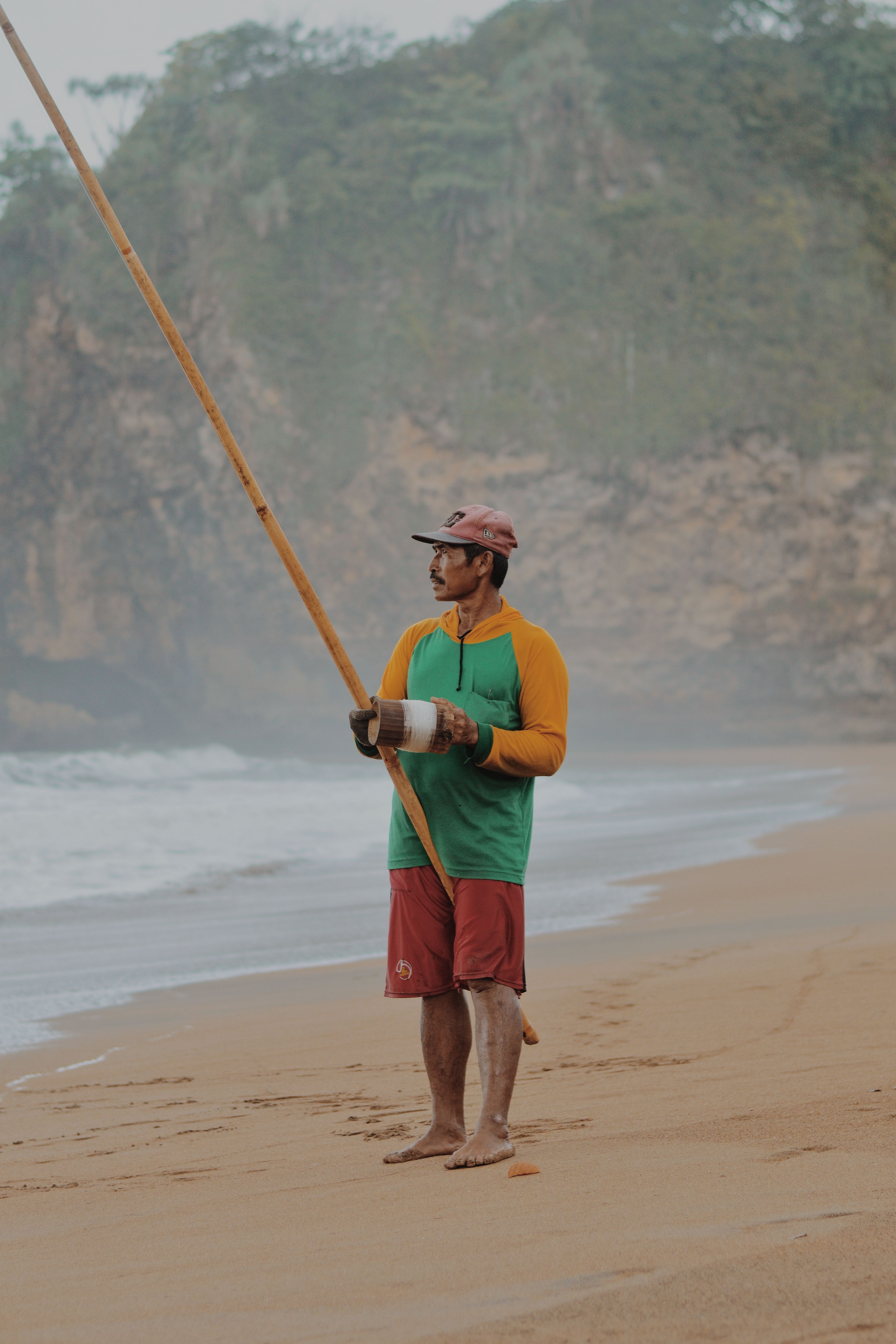 A man in casual attire fishing on a misty beach with a tropical backdrop.