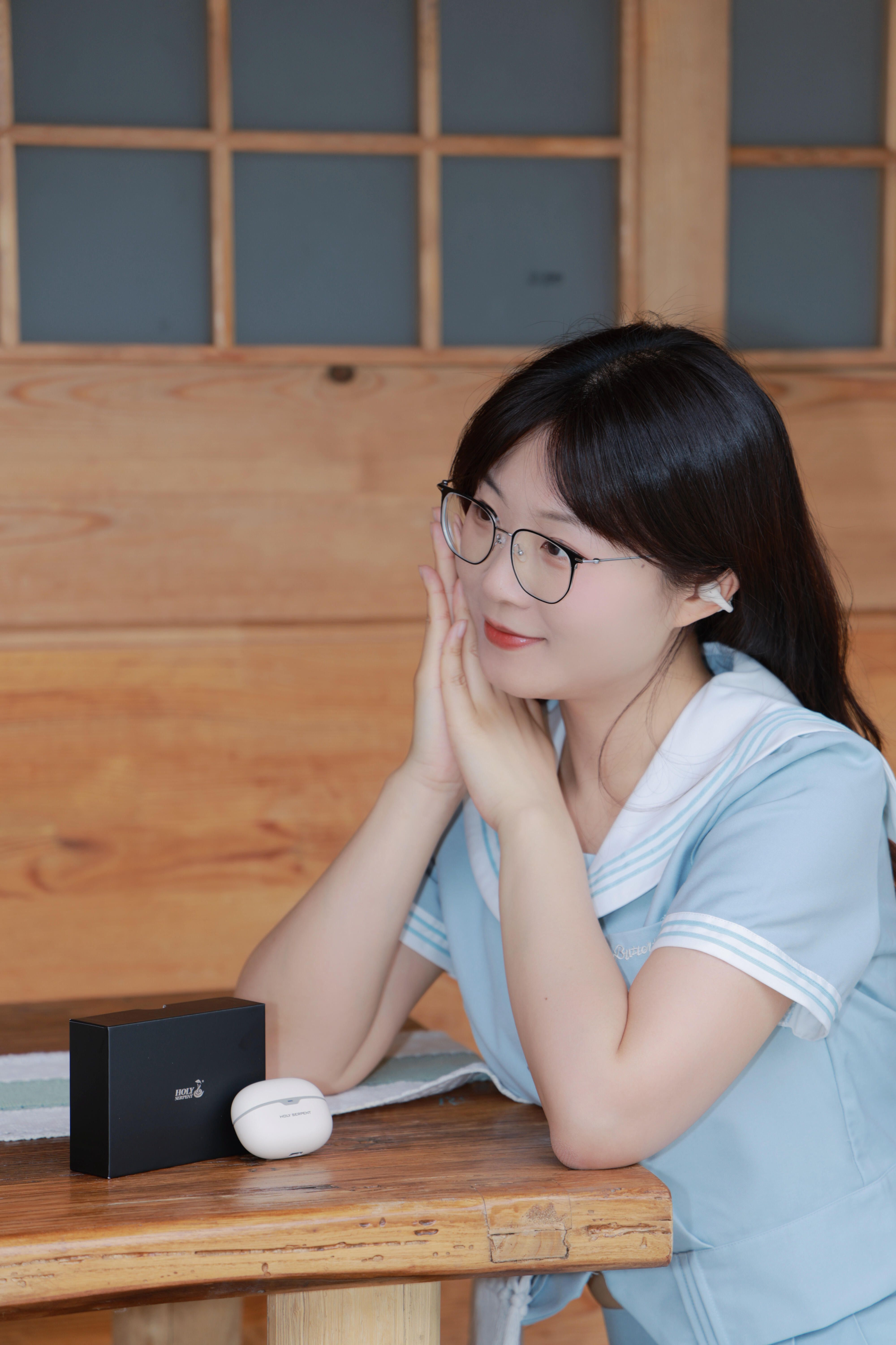 A young woman wearing glasses in a blue uniform sitting at a wooden table indoors, looking thoughtful.