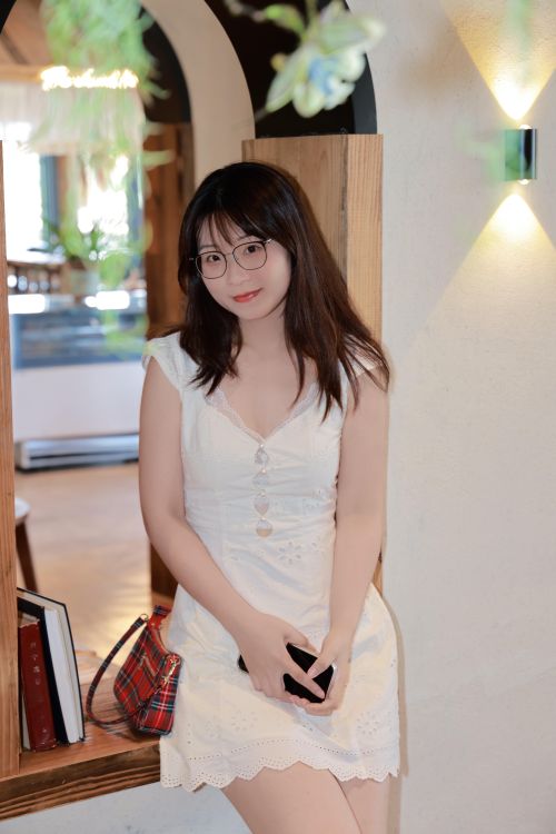 Free A young woman sitting indoors, relaxing in a white dress with eyeglasses and a plaid bag. Stock Photo
