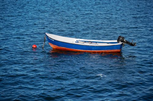 Free A lone blue and orange fishing boat floats on a calm, clear sea with a red buoy nearby. Stock Photo
