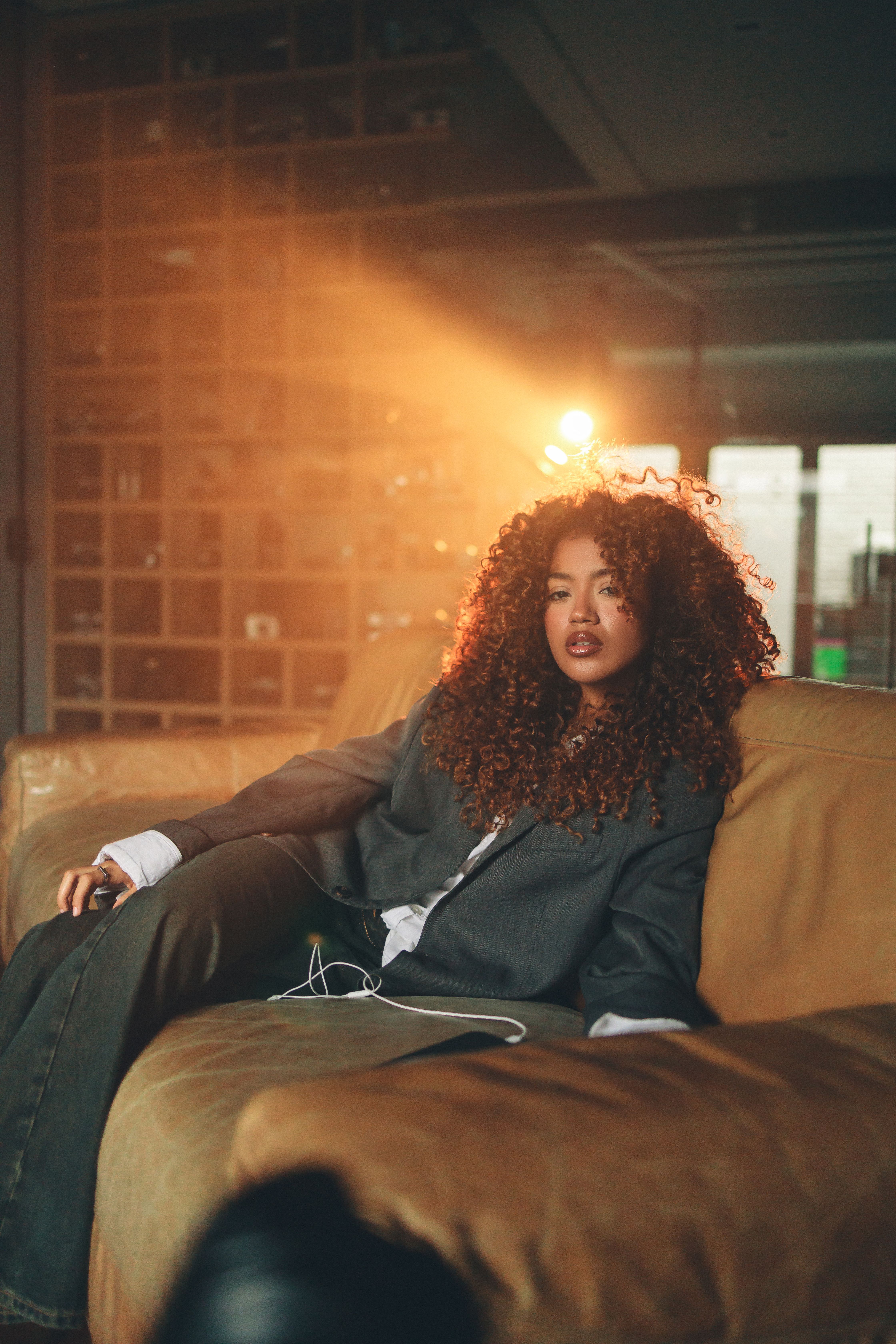 A woman with curly hair relaxes on a couch in warm, moody lighting, indoors.