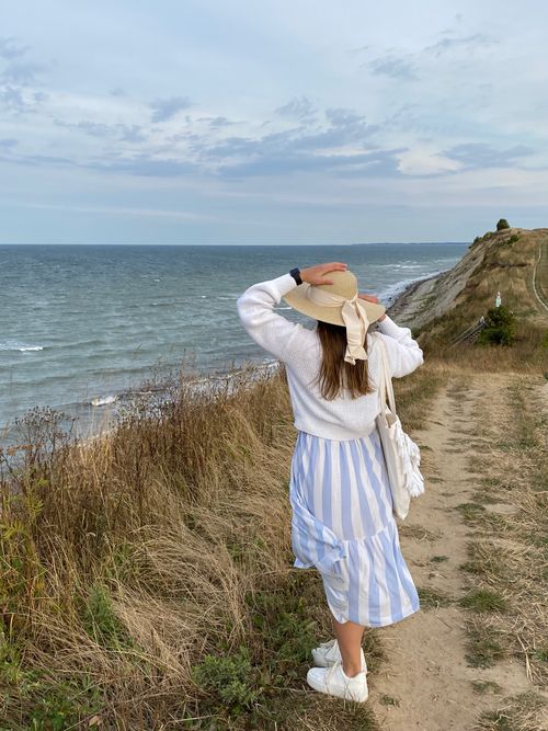 Free A woman views the ocean from a coastal cliff, dressed in a summer outfit and hat. Stock Photo