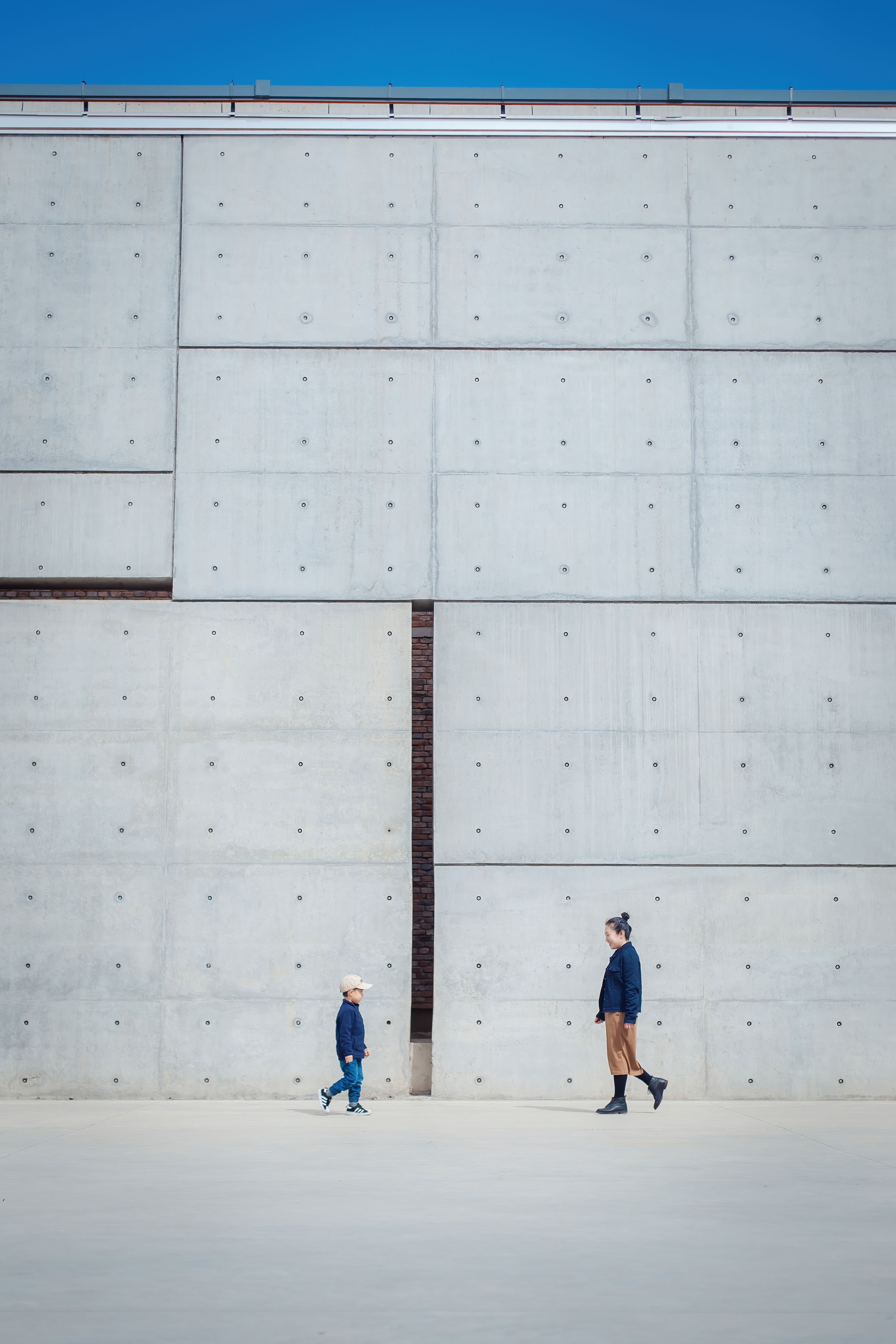 Two individuals walking by a large concrete wall in a minimalist urban setting.