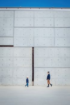 Two individuals walking by a large concrete wall in a minimalist urban setting.