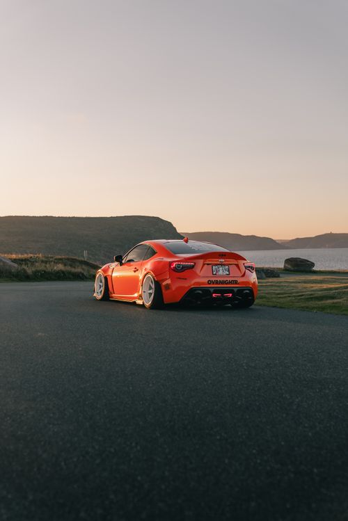 Free Orange sports car drives along a scenic coastal road at sunset with hills in the background. Stock Photo