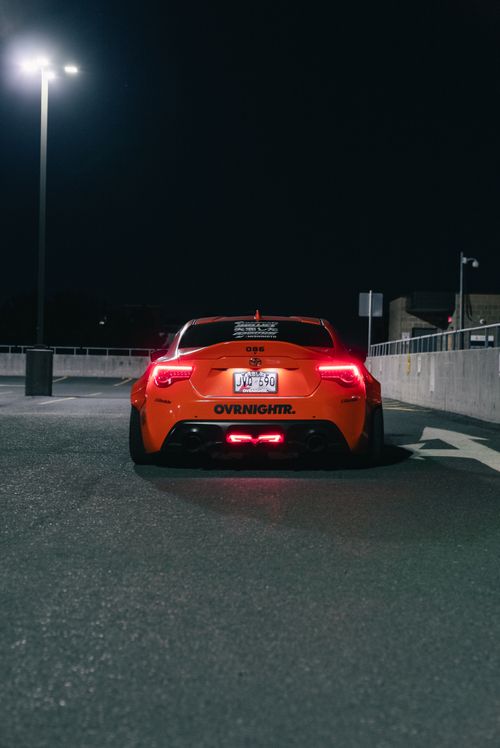 Free Rear view of a bright orange sports car illuminated under street lights at night in a city parking lot. Stock Photo