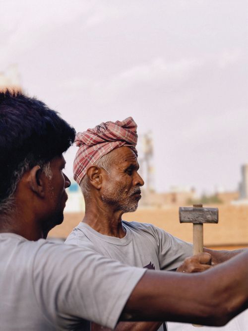 Free An elderly craftsman working with a hammer alongside another person, shot outdoors. Stock Photo