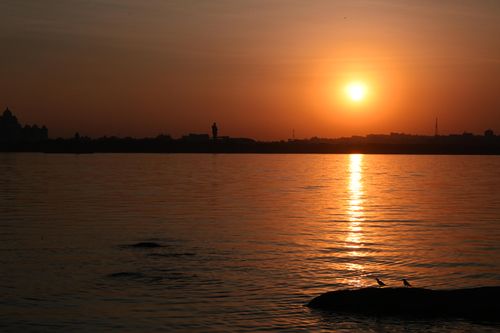 Free A serene sunset over a tranquil lake, featuring silhouetted birds on a rock and a vibrant sky. Stock Photo
