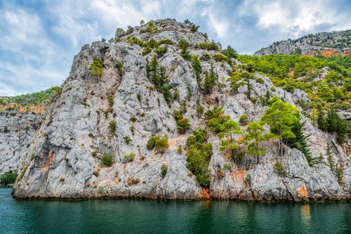 Free Stunning view of a rocky cliff with lush greenery by a calm turquoise sea under a cloudy sky. Stock Photo
