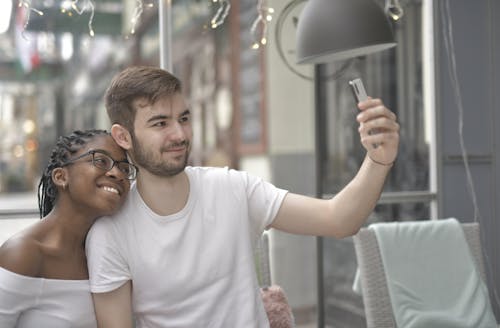 Man in White Crew Neck T-shirt Holding a Smartphone Taking Picture with Her Girlfriend