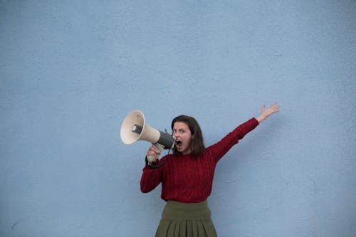 Discontented young speaker in casual wear standing with raised hand and shouting into megaphone while standing near blue concrete wall and looking at camera