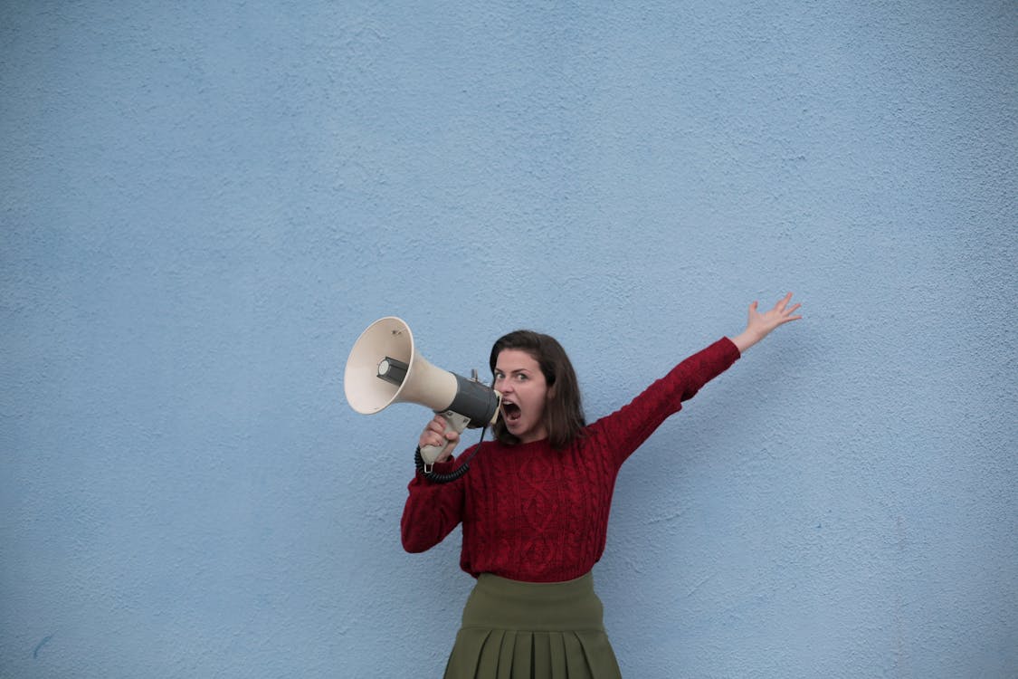 Free Discontented young speaker in casual wear standing with raised hand and shouting into megaphone while standing near blue concrete wall and looking at camera Stock Photo