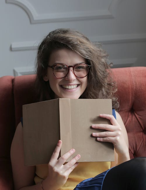 Happy young woman in eyeglasses sitting with book at home