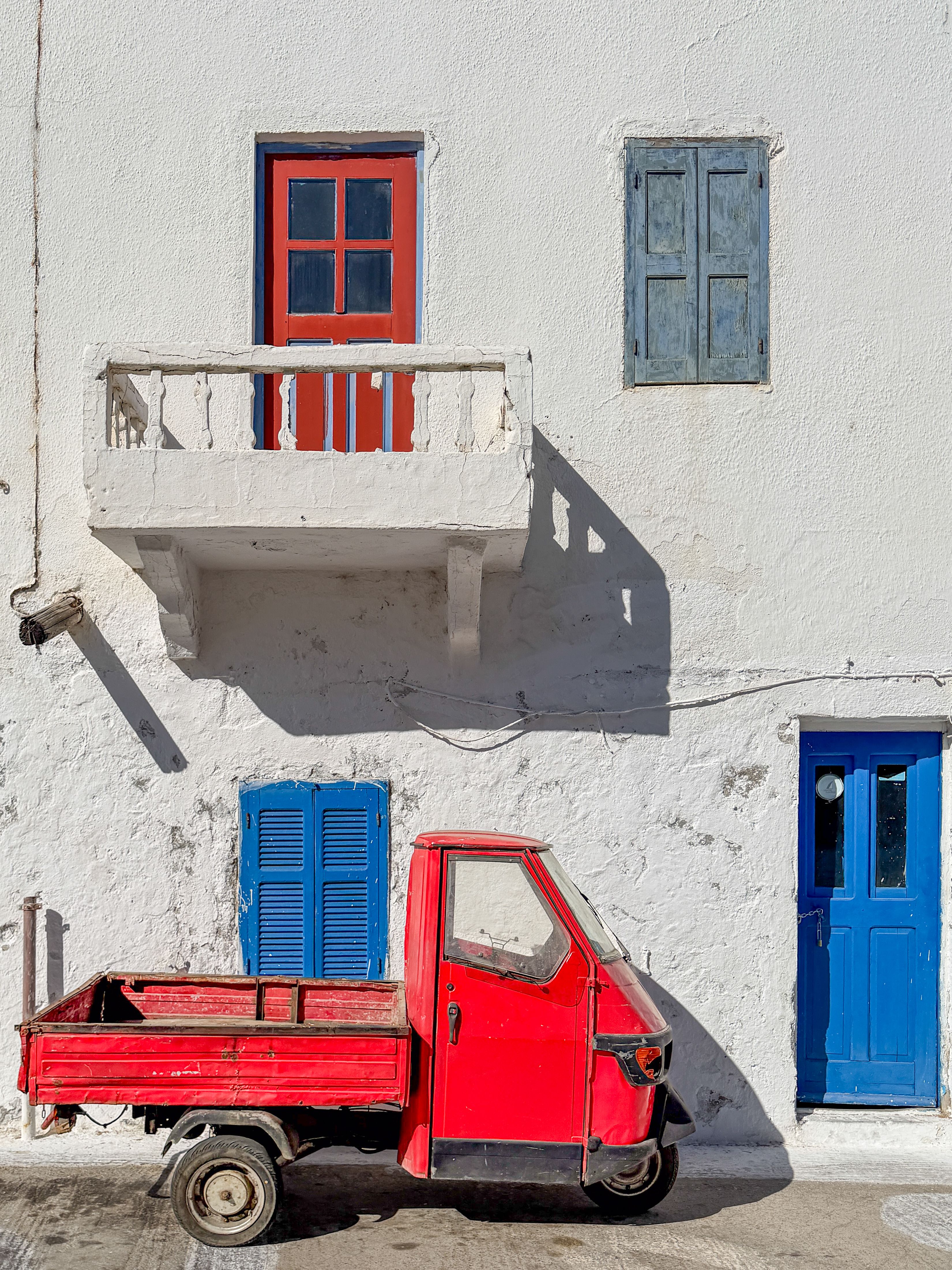 A classic red Vespa parked in front of a vibrant, colorful Greek building. Blue shutters and red balcony accentuate the scene.