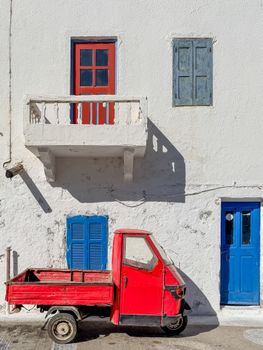 A classic red Vespa parked in front of a vibrant, colorful Greek building. Blue shutters and red balcony accentuate the scene.