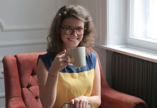 Free Smiling young female in casual clothes and eyeglasses drinking tea while taking book and sitting on cozy chair near window in flat in daylight and looking at camera Stock Photo