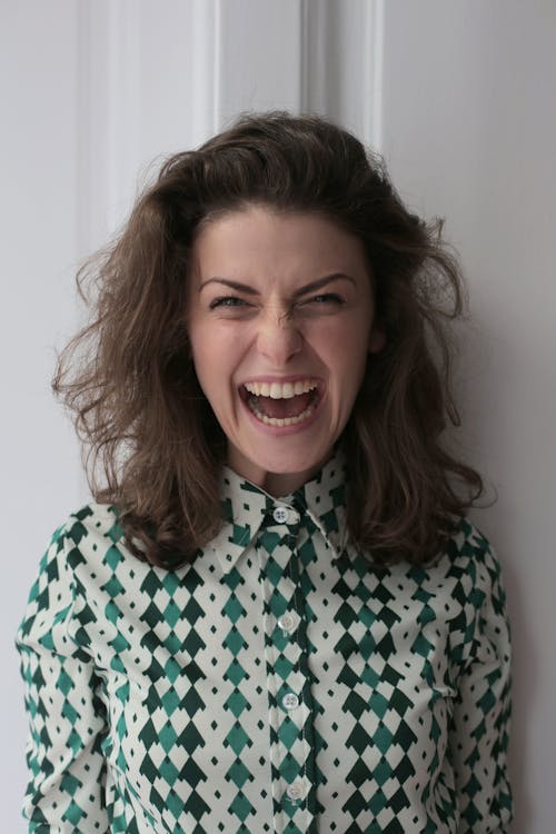 Extremely happy young female in knitted dress standing with open mouth near white wall in apartment and looking at camera