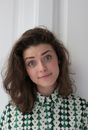 Young female in knitted blouse standing near white wall with tilted head and looking surprisingly at camera in apartment