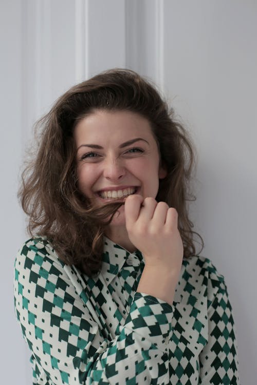 Cheerful young female in knitted dress standing leaned on wall in apartment while touching short wavy hair and looking at camera