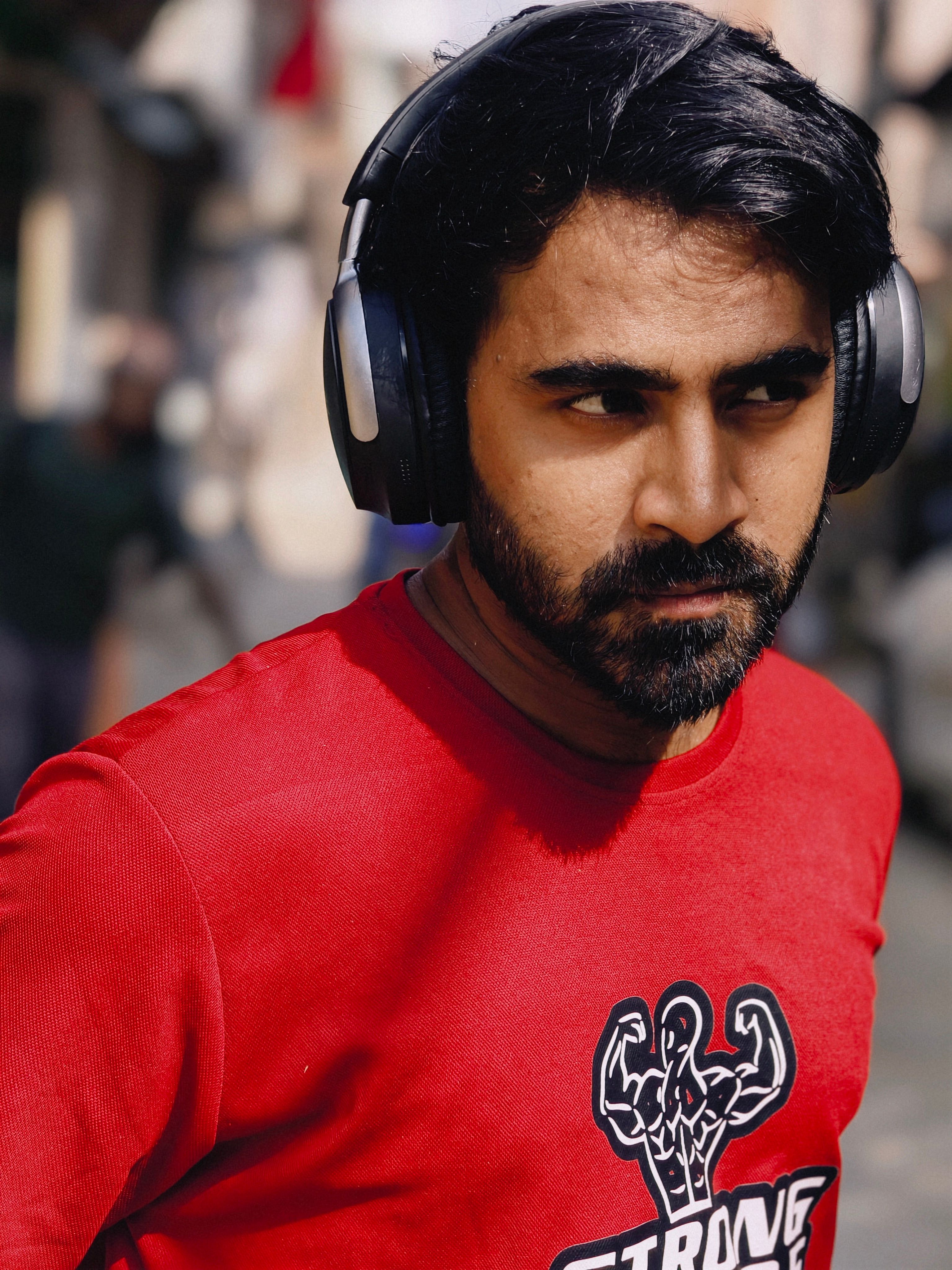 Young man with headphones listening to music outdoors, wearing a red t-shirt on a sunny day.