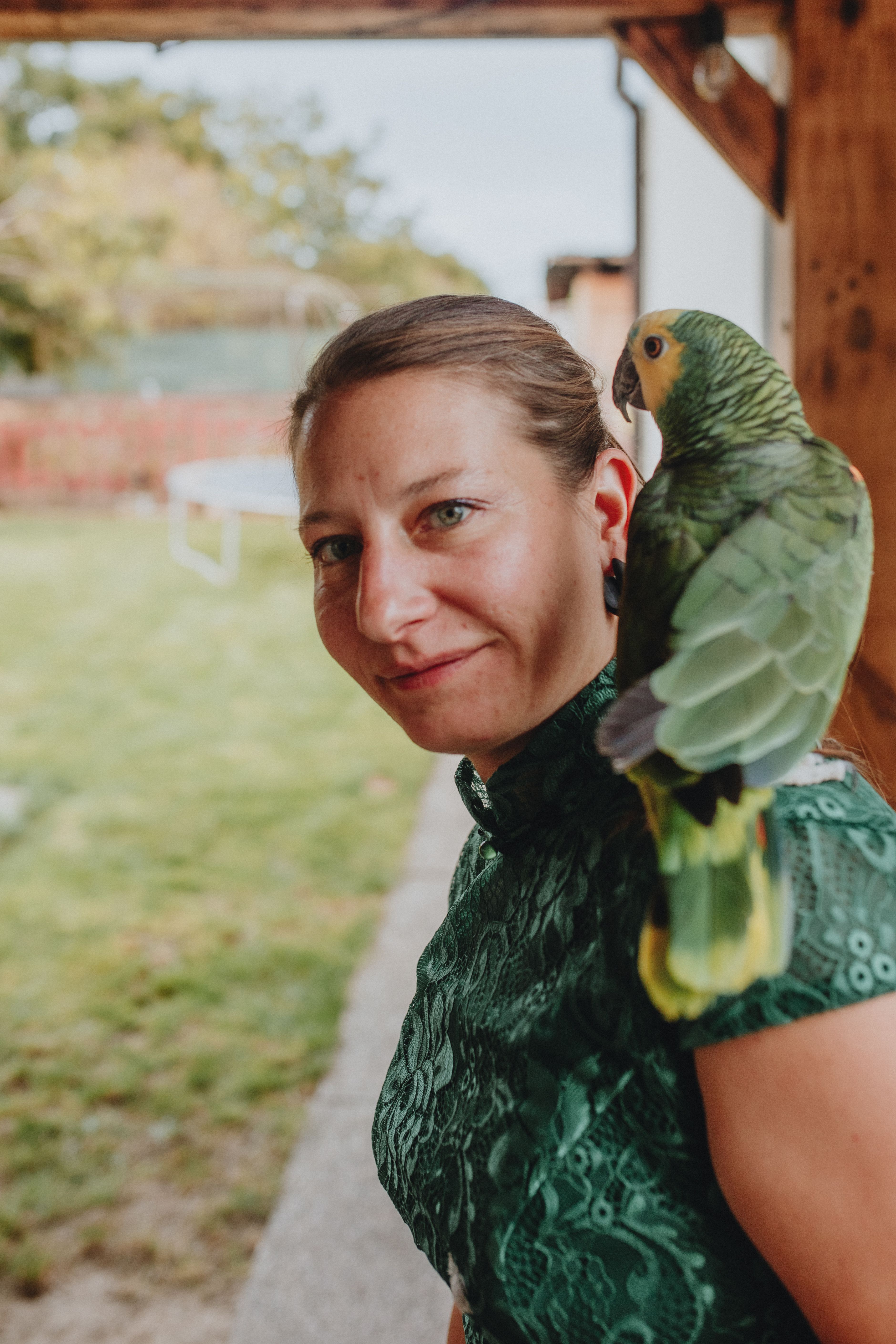 A woman smiling outdoors with a green parrot perched on her shoulder, creating a warm and natural scene.