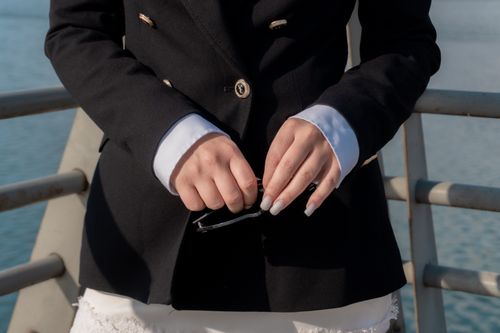 Free Close-up of hands holding sunglasses, wearing a black suit with white cuffs, outdoors by the water. Stock Photo