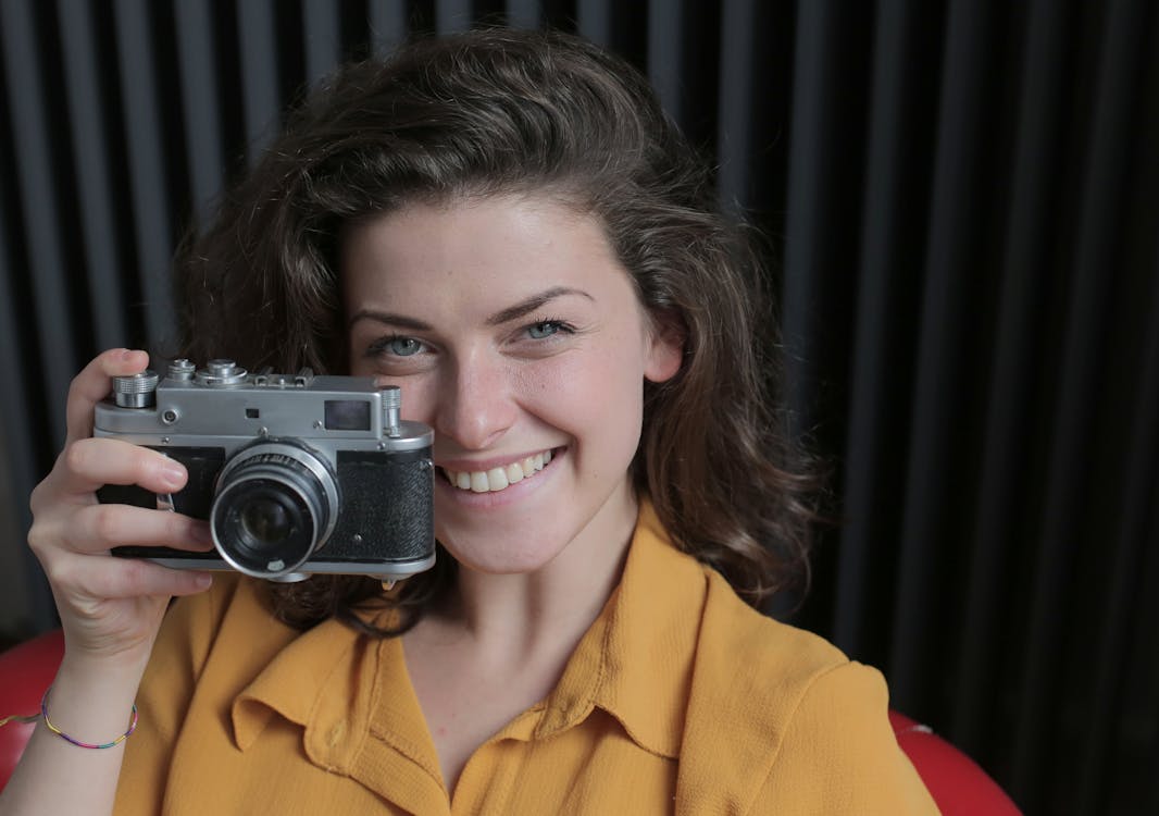 Cheerful young female in casual wear taking photo using digital photo camera while sitting on chair and looking at camera in apartment