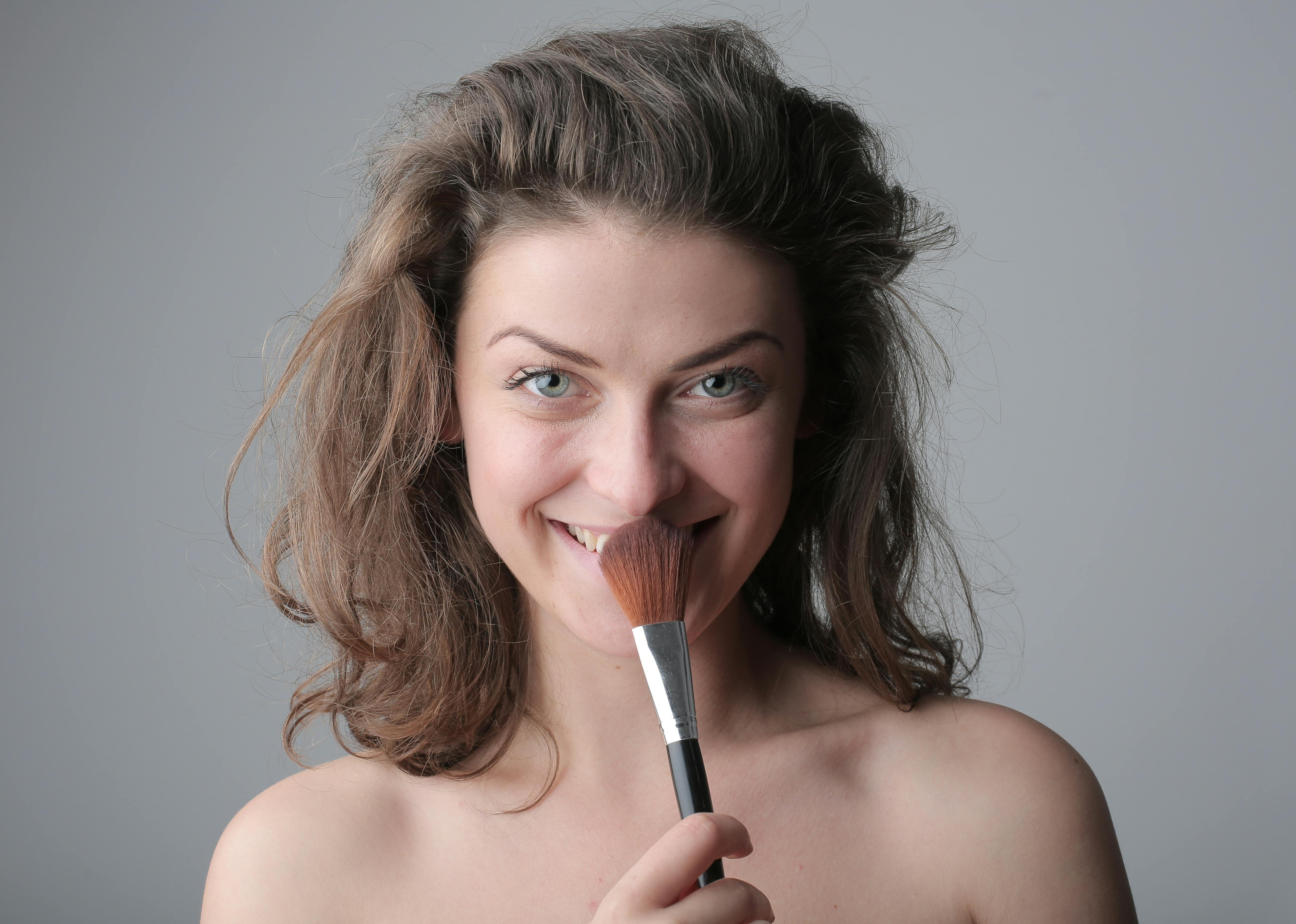 Close-up of a smiling woman holding a makeup brush against gray background.
