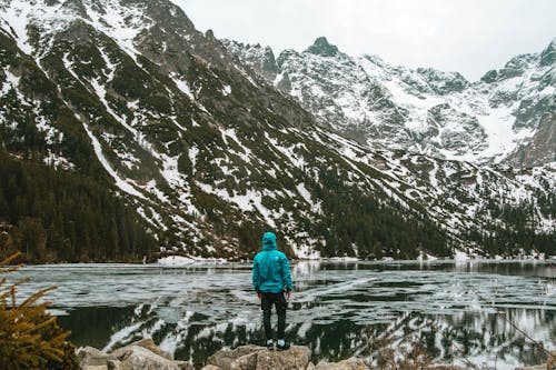 Man in Blue Jacket and Black Pants Standing on Snow Covered Ground Near Snow Covered Mountain