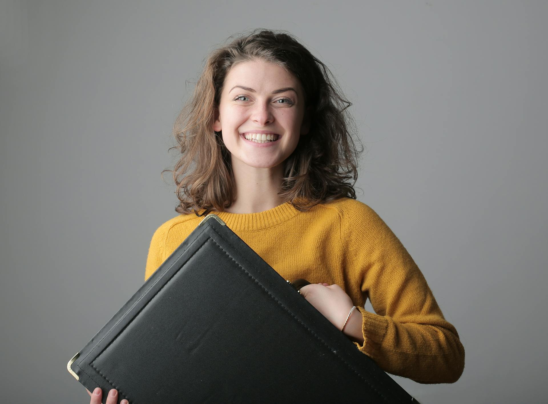 Woman in mustard sweater smiling while holding a black portfolio case against a gray background.