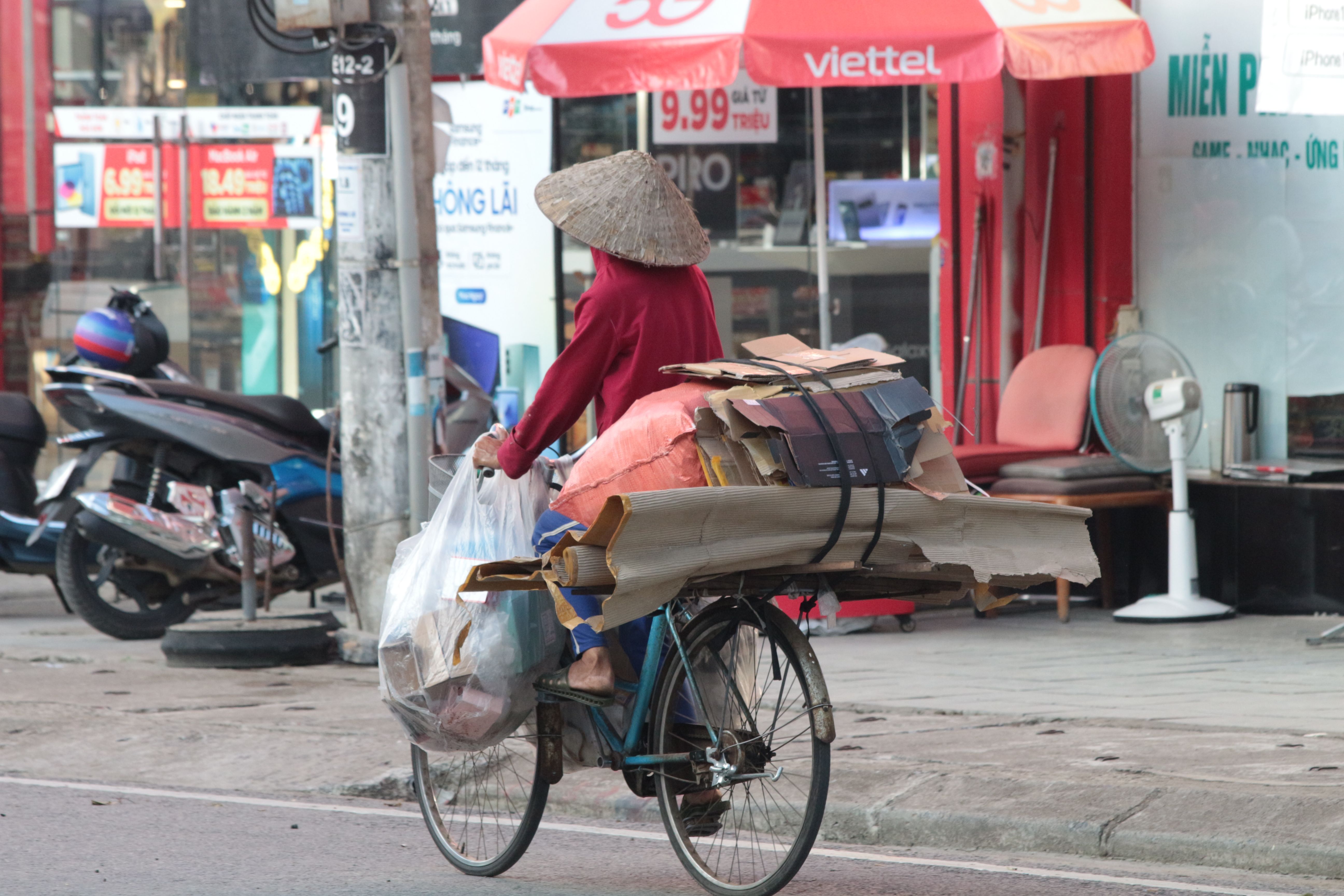 A street vendor in Vietnam rides a bicycle loaded with cardboard and plastic bags.