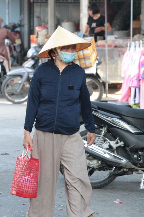 Free Woman wearing a conical hat and mask at a bustling outdoor market with a motorbike visible. Stock Photo