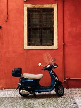 A timeless blue Vespa scooter parked in front of a rustic red wall in the charming streets of Verona, Italy.
