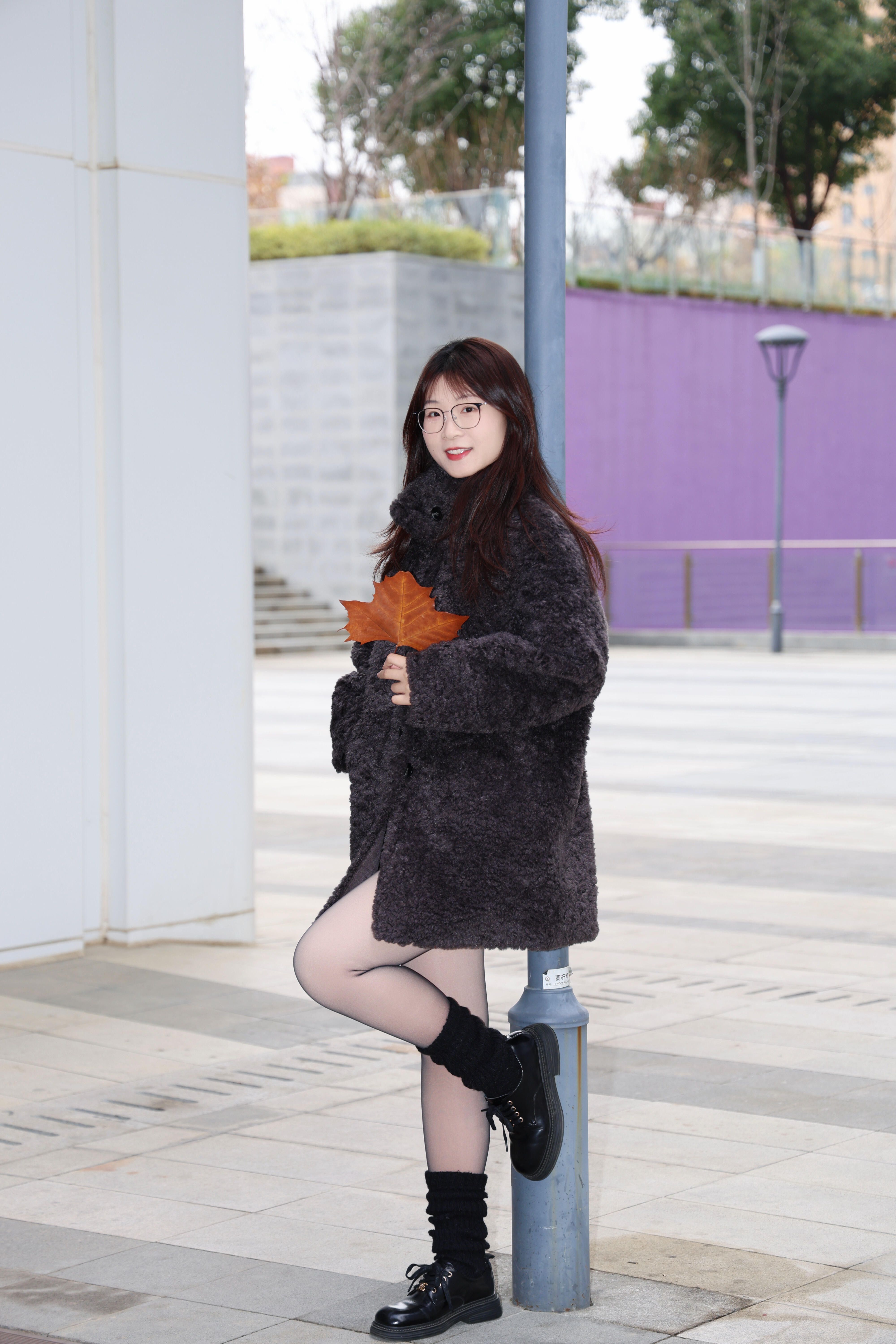Young woman with glasses in a cozy coat holding a fall leaf outdoors.