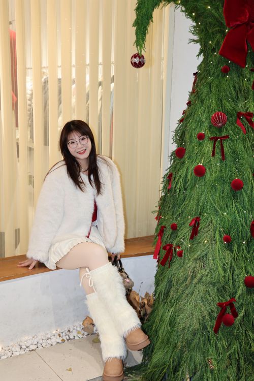 Free A young woman in festive winter attire poses indoors with a decorated Christmas tree. Stock Photo