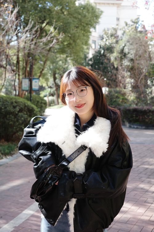 Free A cheerful young woman with glasses smiles in a sunny park, wearing a black coat with white fur trim. Stock Photo