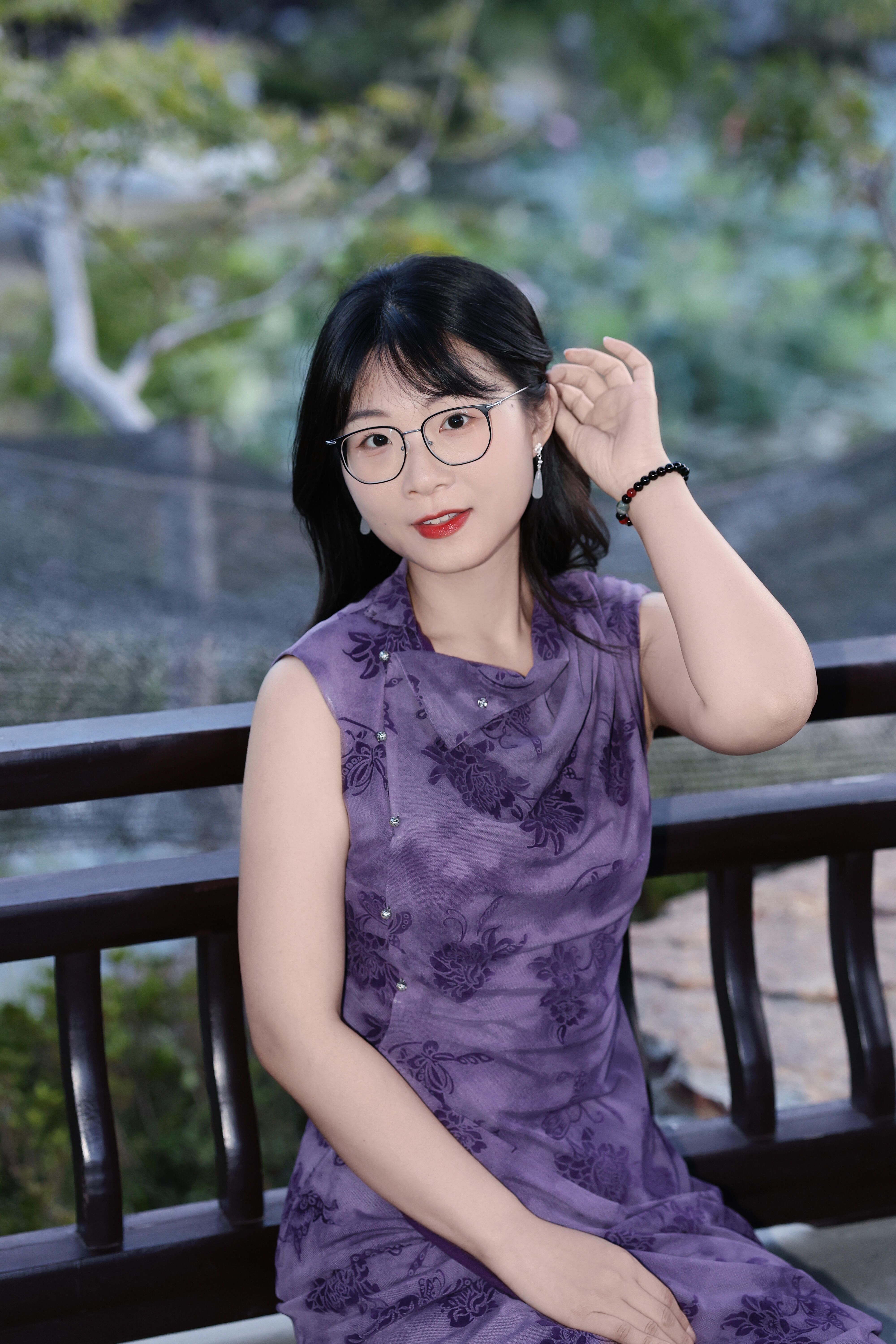 Elegant woman in a floral purple dress wearing glasses, seated on a bench outdoors in a lush garden setting.