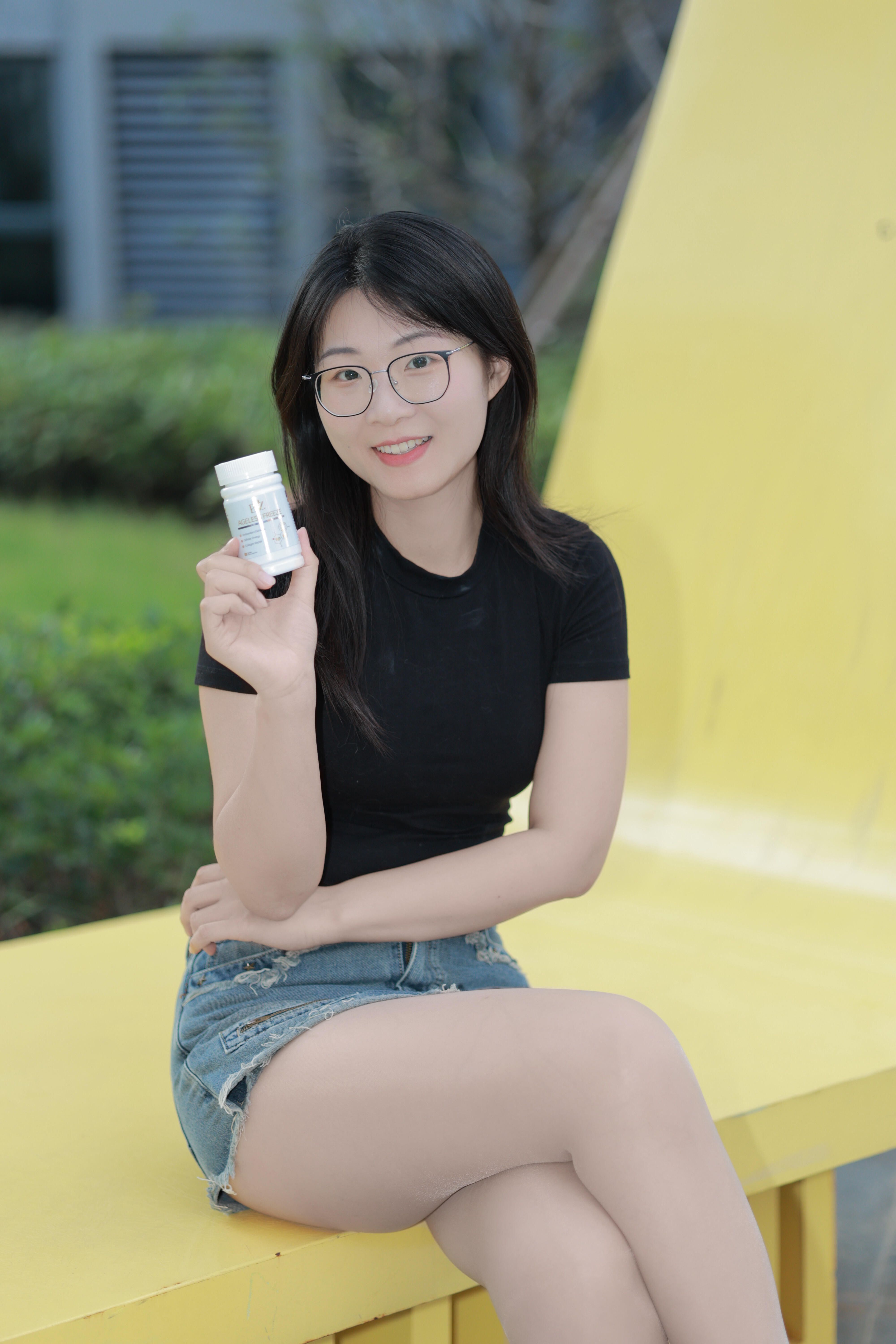 Young woman wearing glasses, seated on a bench outdoors, holding a bottle of supplements, smiling at the camera.