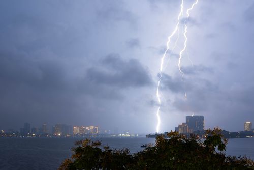 Free A powerful lightning bolt illuminates the night sky above a city skyline, highlighting the silhouette of buildings and the tranquil lake below. Stock Photo