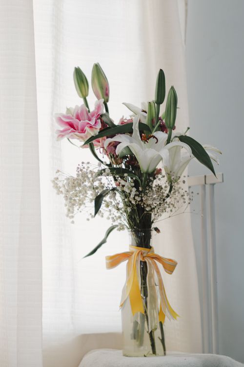 Free A delicate floral arrangement of lilies in a vase, adorned with a yellow ribbon, against a soft curtain backdrop. Stock Photo