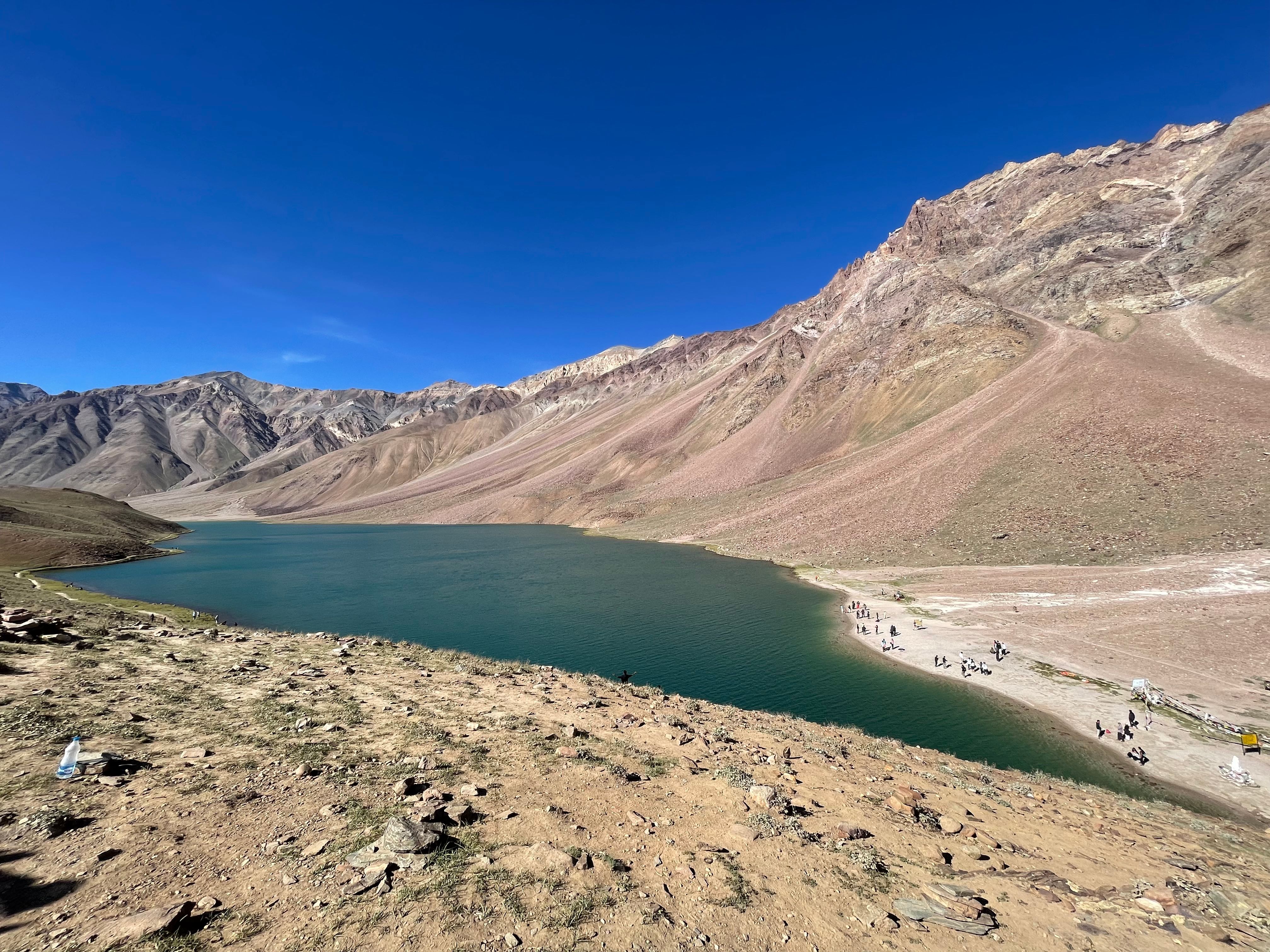 Panoramic view of Chandra Taal Lake surrounded by barren mountains under a clear blue sky in Spiti Valley.