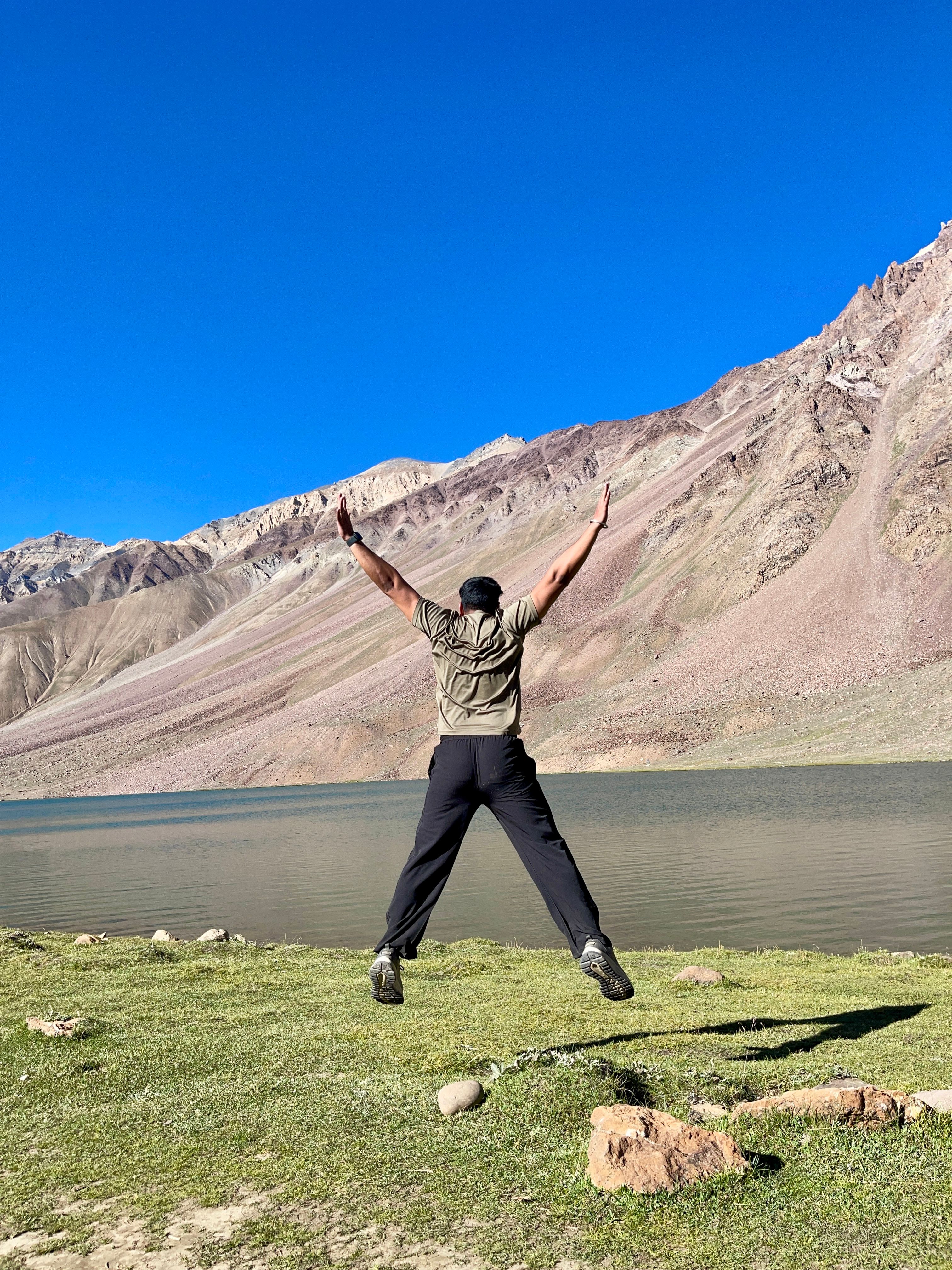 Person jumping in excitement by a serene Himalayan lake under a clear blue sky. Captivating outdoor adventure.