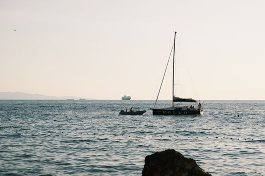 Sailing boat at sunset with distant cargo ships and calm sea.
