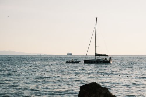 Free Sailing boat at sunset with distant cargo ships and calm sea. Stock Photo