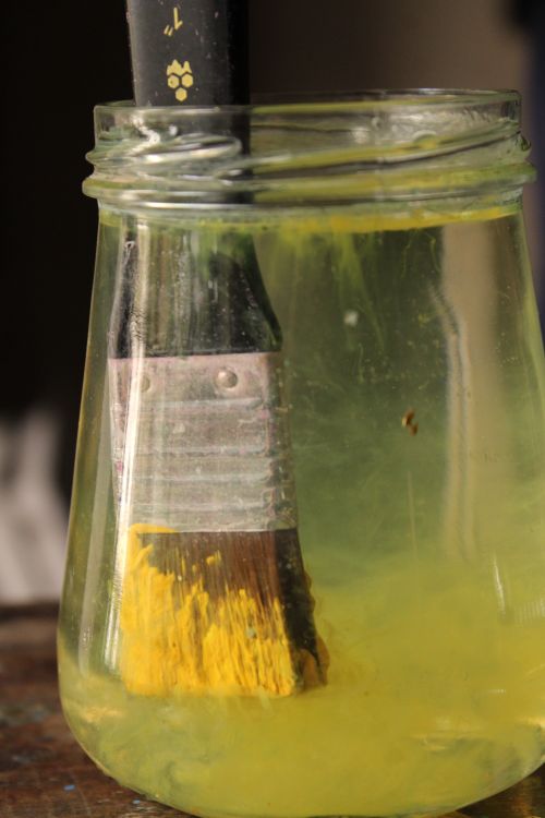 Free A paintbrush resting in a glass jar filled with yellow paint water. Stock Photo