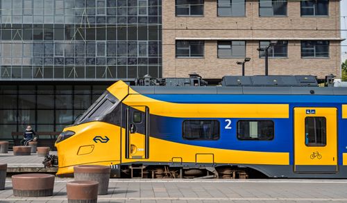 Free Yellow and blue train in urban station with building backdrop and passenger waiting. Stock Photo