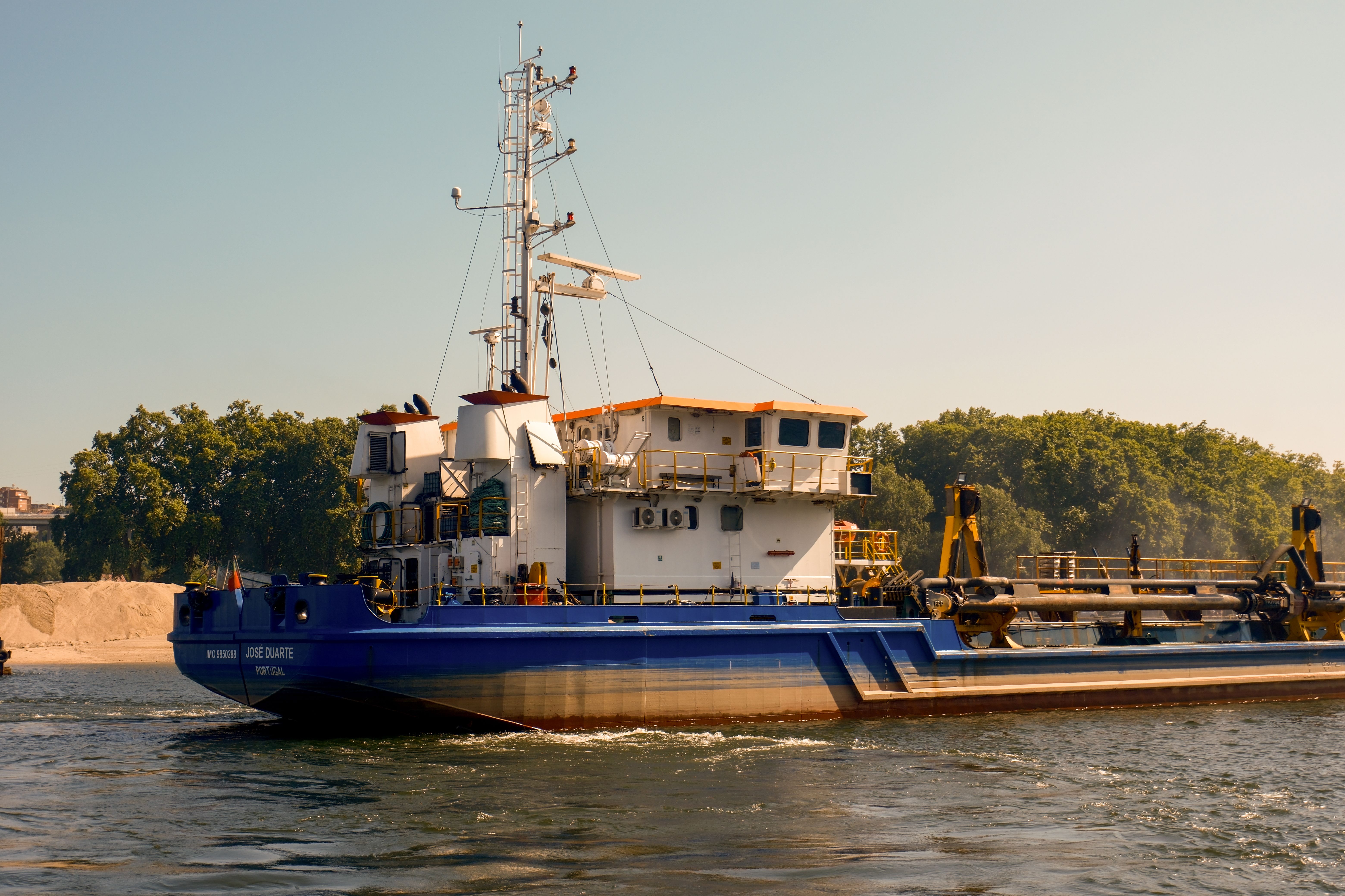 A cargo ship navigating a river with lush greenery in the background on a sunny day.