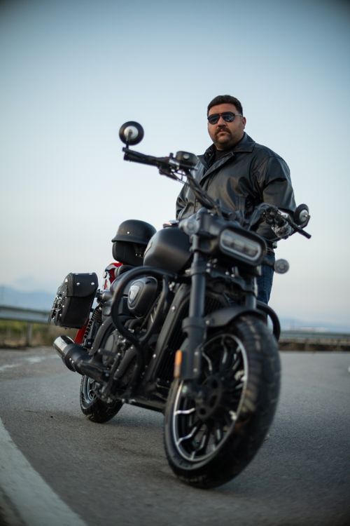 Free A man in a leather jacket and sunglasses leans on a black motorcycle on a road. Stock Photo