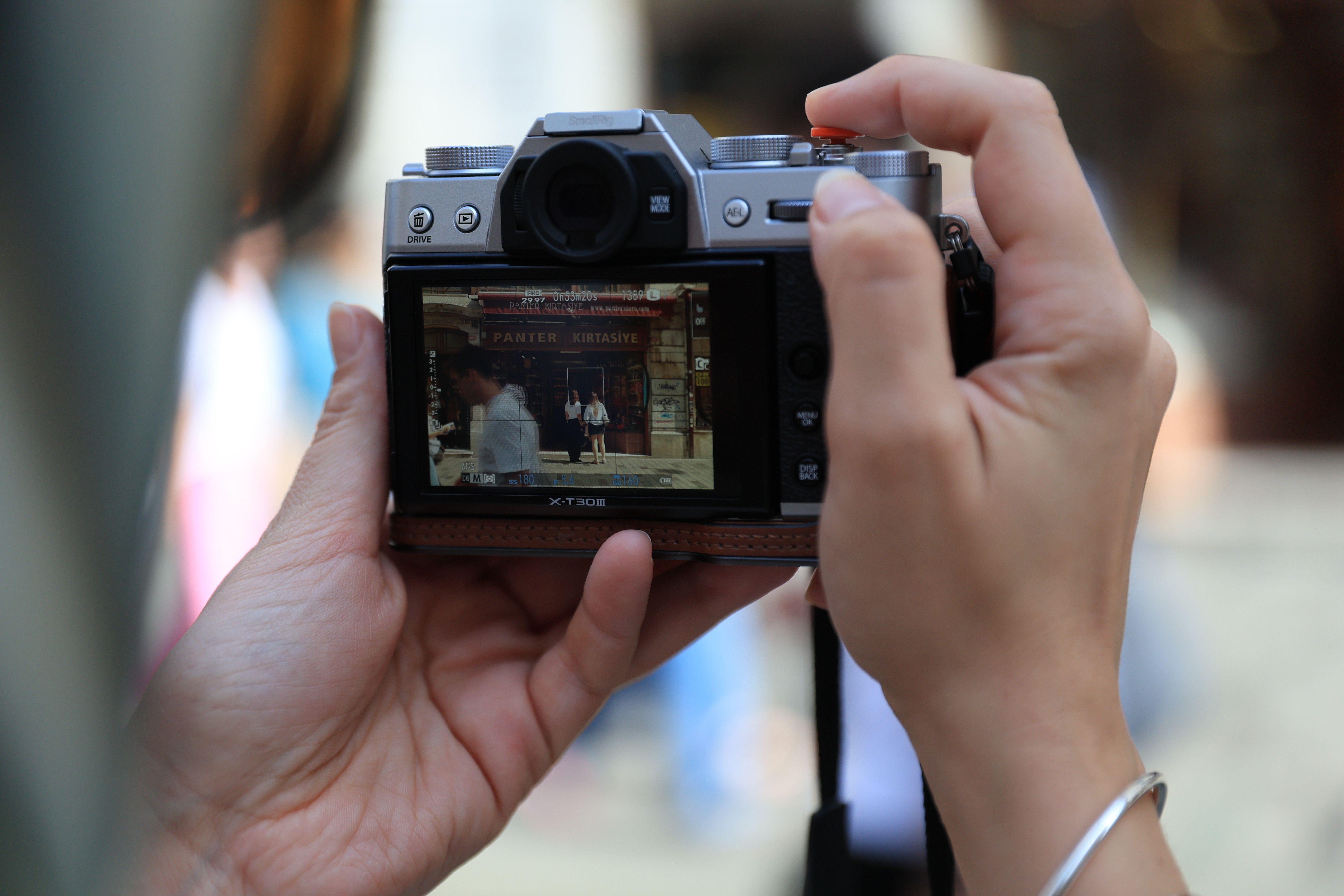 Hands hold a camera capturing a busy outdoor street scene in soft daylight.