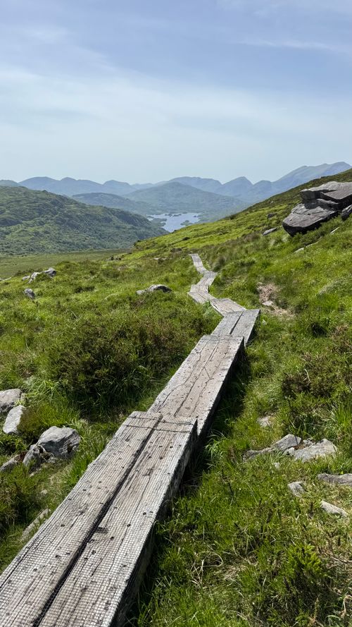 Free Wooden path on lush green mountainside overlooking scenic valley and distant hills. Stock Photo